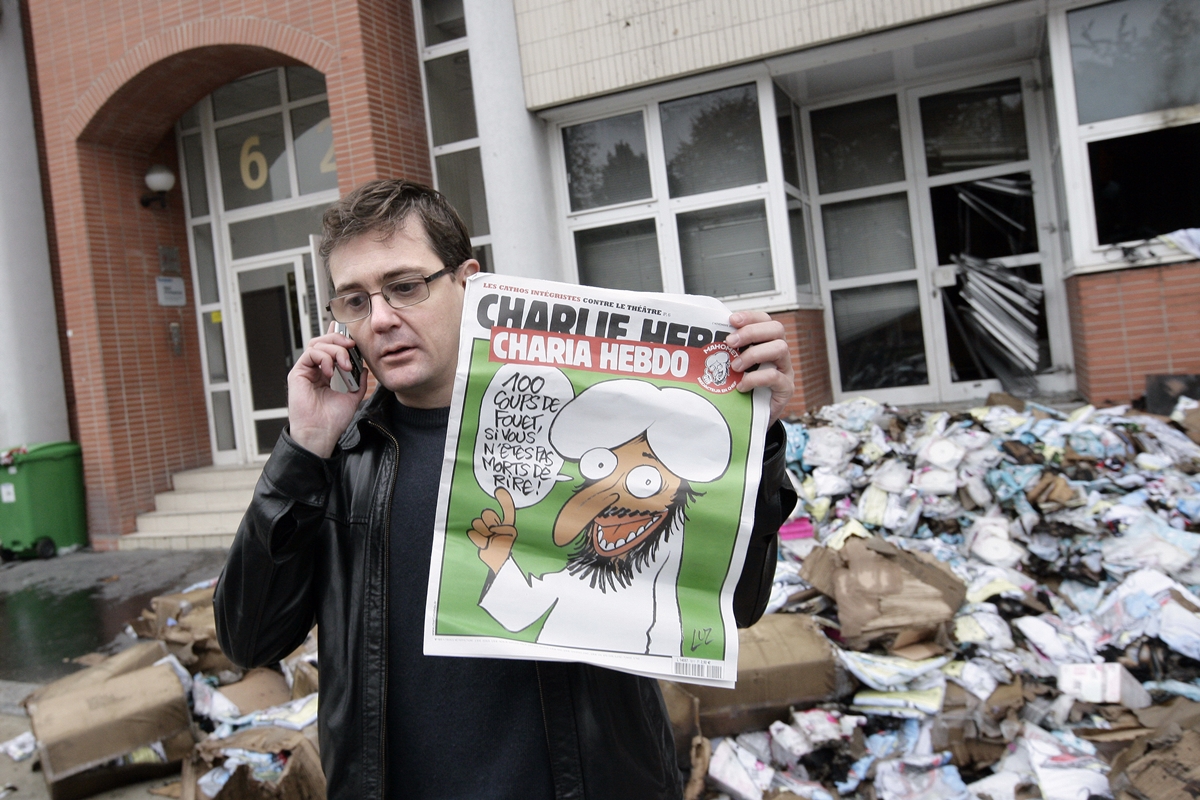 Paris 2. november 2011: Sjefredaktør i Charlie Hebdo Stéphane Charbonnier (Charb) holder en utgave av bladet med Muhammed-karikatur på forsiden. Tekst i snakkeboblen: "100 piskeslag om du ikke dør av latter". Redaksjonslokalene i bakgrunnen ble ødelagt av en brannbombe natten før. (Foto: Alexander Klein, AFP/NTB Scanpix).